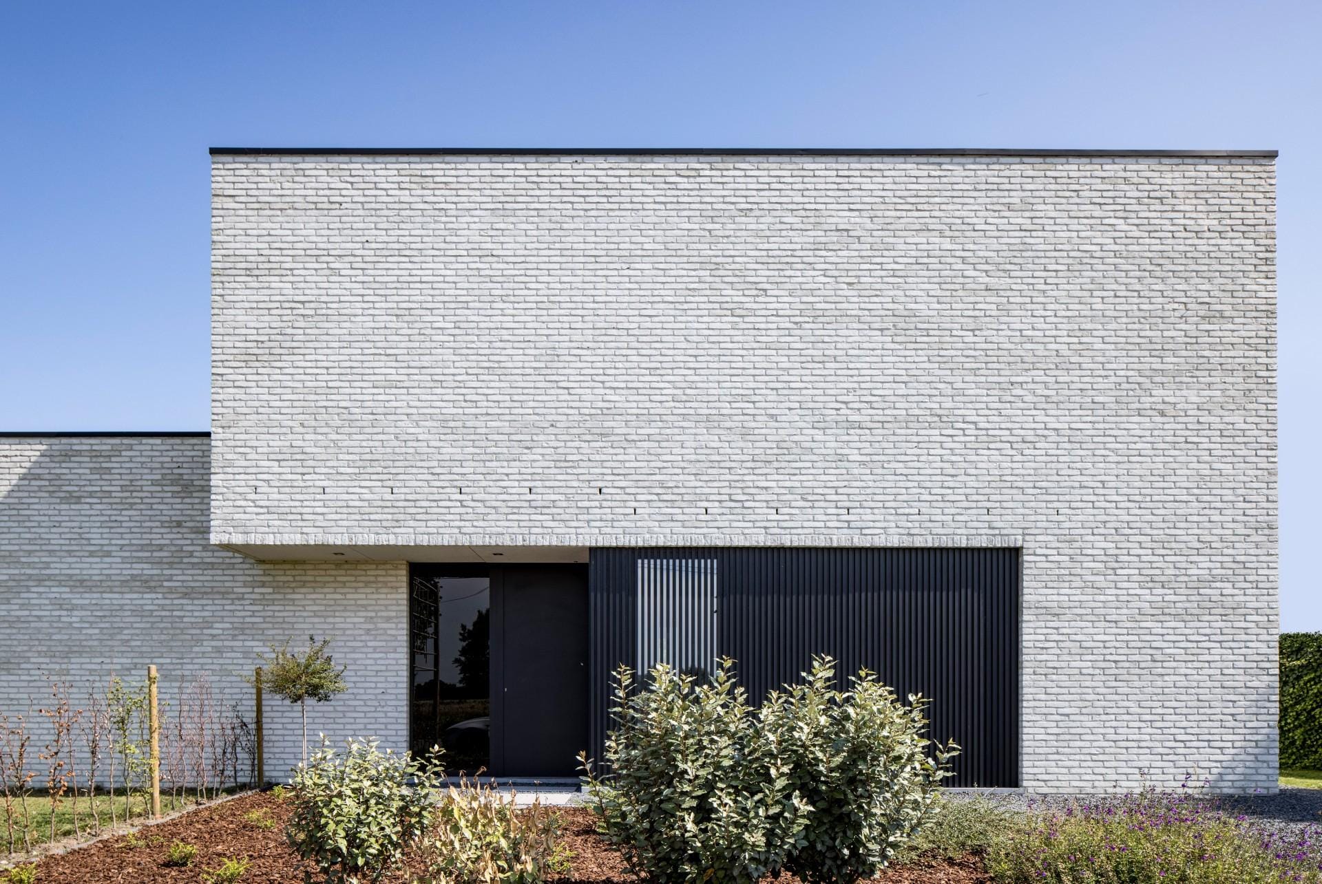 Sobere moderne woning met witte gevelsteen, donkere voordeur en houten lamellenpartij.